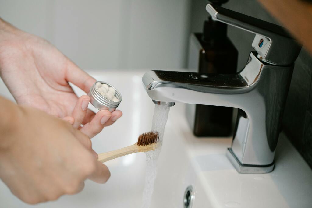 Une brosse à dent est tendue sous le robinet d'eau. Dans l'autre main, une personne tient une boîte avec du dentifrice solide en pastilles.